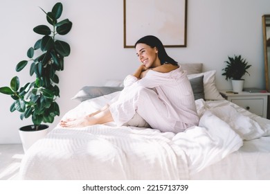 Side view of smiling young lady in nightwear sitting on bed resting on weekend in modern bedroom and looking away - Powered by Shutterstock