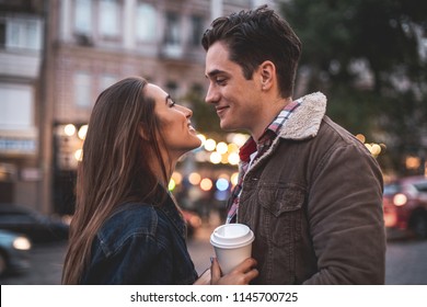 Side view of smiling young male and female looking at each other with delight. They are standing outside and holding coffee cups in hands