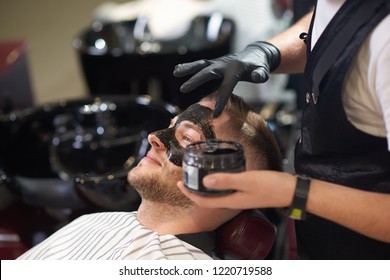 Side View Of Smiling Young Man Lying On Chair In Vintage Barber Shop While Barber In Protective Gloves Putting Black Mask Against Dots On Face. Concept Of Skin Care And Cleaning.