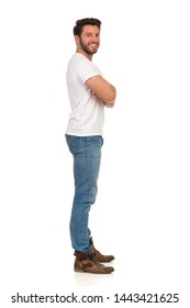 Side View Of Smiling Young Man In Jeans And White T-shirt Standing With Arms Crossed And Looking At Camera. Full Length Studio Shot Isolated On White.