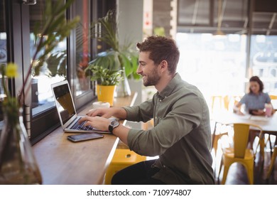 Side View Of Smiling Young Man Typing On Laptop At Counter In Coffee Shop