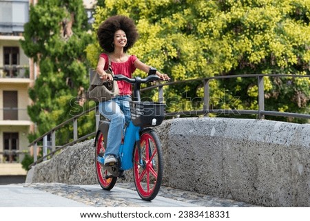 Side view of smiling young Moroccan female with handbag looking away while riding electric bicycle on city bridge with railing against blurred green trees and building in daylight