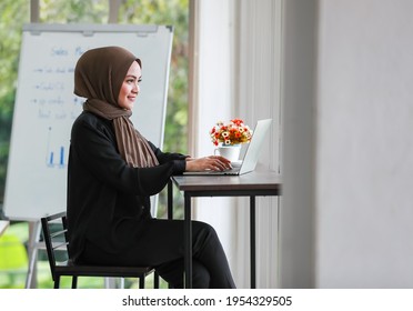 Side View Of Smiling Young Muslim Female In Traditional Clothes And Hijab Sitting At Table With Laptop In Coffee Breaking Time In Modern Workplace