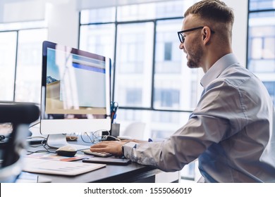Side view of smiling young office worker in shirt and eyeglasses using computer at office desk smiling with successful results  - Shutterstock ID 1555066019