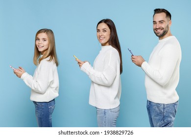 Side View Of Smiling Young Parents Mom Dad With Child Kid Daughter Teen Girl Looking Camera Using Mobile Phone Typing Sms Message Isolated On Blue Background Studio. Family Day Parenthood Concept