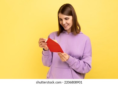 Side View Of Smiling Young Woman Opening Envelope And Reading Letter, With Positive Expression, Got Unexpected Pleasant News, Wearing Purple Hoodie. Indoor Studio Shot Isolated On Yellow Background.