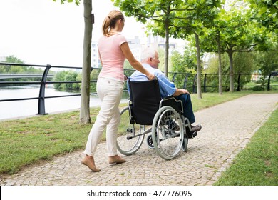 Side View Of A Smiling Young Woman Assisting Her Disabled Father On Wheelchair In Park