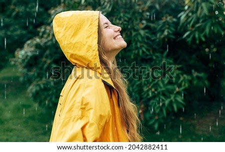 Side view of a smiling young woman wearing a yellow raincoat during the rain in the park. Cheerful female enjoying the rain outdoors. A woman has a joyful expression during enjoying the rainy weather.