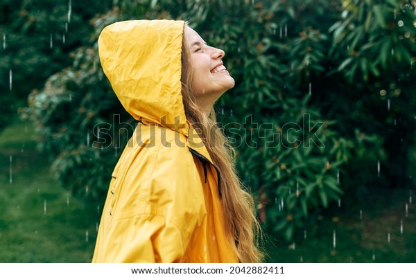 Side view of a smiling young woman wearing a\
yellow raincoat during the rain in the park. Cheerful female\
enjoying the rain outdoors. A woman has a joyful expression during\
enjoying the rainy\
weather.