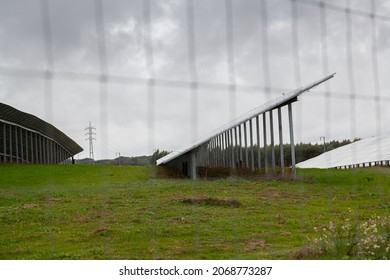 Side view of solar panels set at angle in green and brown autumn field behind wire fence under grey sky in rainy day in Portugal
