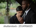 Side view of sorrowful Black man comforting grieving senior woman leaning on his shoulder at memorial service. Mourning couple standing under umbrella at cemetery, copy space