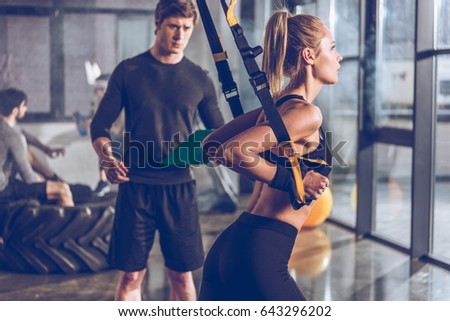 side view of sportive woman exercising with trx gym equipment with trainer near by