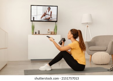 Side View Of Sporty African American Female Watching Sport Channel On TV, Hydrating And Exercising Sitting On Mat At Home. Side View. Healthy Weight Loss And Workout, Fitness Lifestyle Concept