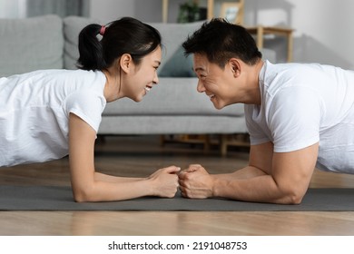 Side View Of Sporty Asian Man And Woman In White Sportswear Planking Together, Standing On Elbows Face To Face And Smiling, Encourage Each Other While Having Workout At Home, Closeup