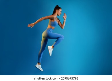 Side View Of Sporty Woman Exercising Running Or Doing Elbow-To-Knee Crunch Standing Posing In Mid-Air On Blue Studio Background, Looking Aside. Sport And Fitness Motivation. Full Length