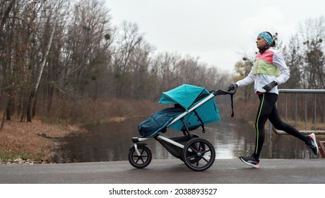 Side view of sporty young woman mom running with a stroller along an empty park on a cold autumn day. Parenting, healthy lifestyle, sports concept
