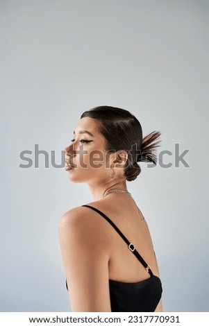 side view of spring style asian woman with bold makeup, brunette hair, trendy hairstyle, in silver necklaces, black and elegant strap dress on grey background, spring fashionable photography