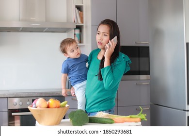 Side View Stressed And Busy Mother Talking On Phone With One Hand Holding Her Baby In Kitchen Home. Mixed Race Asian-German Family Mom And Son. Housewife Overwork Anxiety.