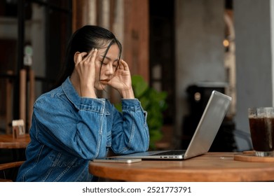 A side view of a stressed, thoughtful Asian woman in a denim jacket, feeling headache and tired, facing a problem with her project while working on her laptop remotely from a coffee shop. - Powered by Shutterstock