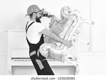 Side View Strong Bearded Man Carrying Old Armchair In Front Of Piano. Mover Walking With Fragile Old Piece Of Furniture In His Hands. Moving To New Place.