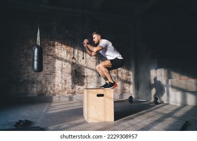 Side view of strong male athlete in sportswear jumping on to wooden box from floor with raising hands in evening light in gym - Powered by Shutterstock