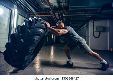 Side view of strong muscular Caucasian bodybuilder flipping massive tire in hallway. Night workout concept.