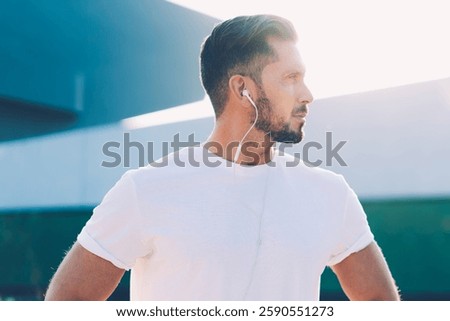 Side view of strong powerful runner with earphones listening music and looking along with hands aside in bright day along glass building