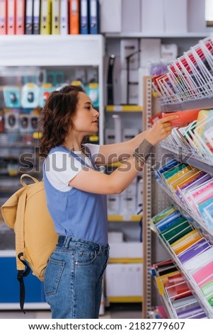 side view of student with backpack choosing copybook on rack with stationery