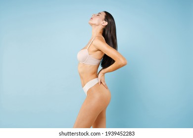 Side view of stunning seductive young brunette woman 20s in beige underwear showing fit sexy body standing posing put hands on back isolated on pastel blue colour wall background studio portrait