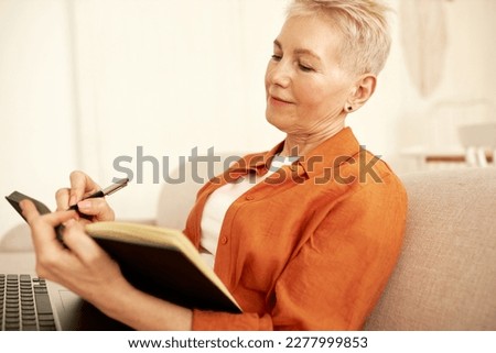 Side view of stylish aged female novelist sitting on couch with laptop on knees noting down thoughts and ideas for new book. Mature woman writing in her diary while sitting on sofa