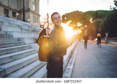 Side View Of Stylish Female Student With Backpack Holding Mobile Phone Laughing At Camera Against Street Steps And Park In Bright Back Lit