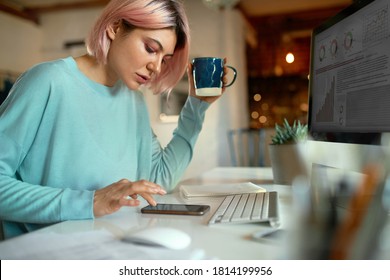 Side View Of Stylish Pink Haired Young Female Blogger Sitting At Table In Front Of Desktop Computer, Using Smart Phone Checking Comments Under Her Post, Drinking Morning Coffee. Electronic Gadgets