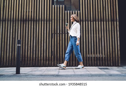 Side view of stylish positive middle aged female in jeans and sneakers strolling along street near modern building and drinking coffee - Shutterstock ID 1935848515