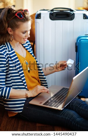 Side view of stylish woman in striped jacket with credit card booking flights online on a laptop while sitting near couch and trolley bags in the modern house in sunny hot summer day.