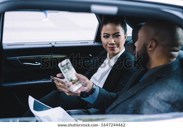 Side view of successful woman\
browsing smartphone listening to African American partner\
discussing strategy of project using mobile sitting on back seat in\
car