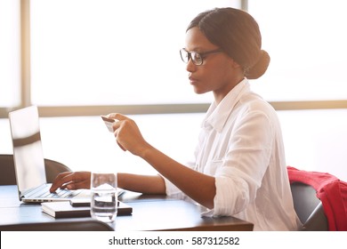 Side View Of Successful Young Adult Black Woman Making Electronic Payments Online With Her Computer And Credit Card With Large Bright Windows Behind Her.