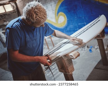 Side view of tattooed male shaper master cutting rail tape for gluing on work piece of surfboard while working in professional joinery workshop - Powered by Shutterstock