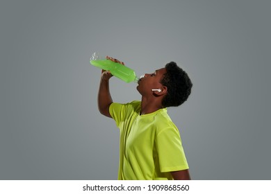 Side View Of A Teenage African Boy Wearing Sports Clothes Drinking Energy Drink While Standing Against Grey Background. Young People And Sport Concept
