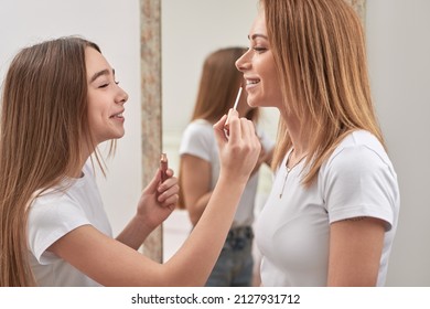 Side view of teenage girl applying lip gloss on lips of mother while doing makeup together in room at home