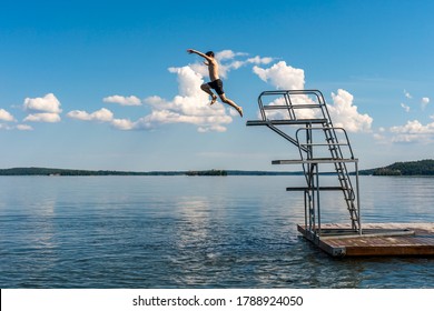 Side View Of A Teenage Male Jump Diving From A Diving Tower With Blue Sky And Horizon In The Background.