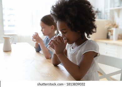 Side View Thirsty Energetic Little Cute Mixed Race Children Girls Drinking Fresh Cool Stilled Water From Glasses, Sitting At Wooden Table In Kitchen, Copy Space For Healthcare Habit Advertisement.