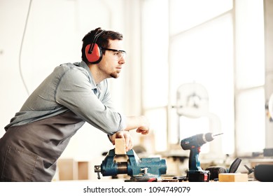 Side view of thoughtful carpenter wearing protective eyewear and earmuffs and leaning on workbench vice with piece of wood in it - Powered by Shutterstock