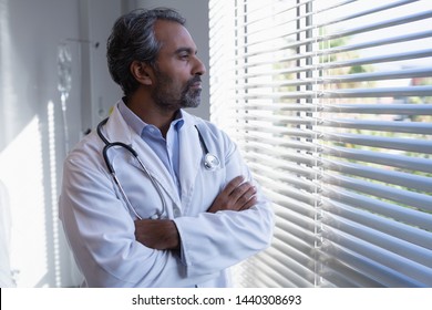 Side View Of Thoughtful Mature Mixed Race Male Doctor Standing With Arm Crossed And Looking Through Window In Clinic At Hospital