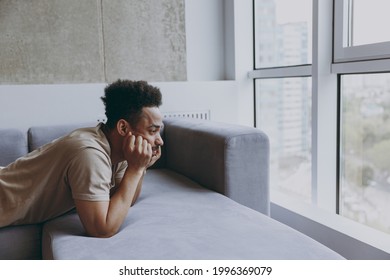 Side view thoughtful pensive sad young african american man 20s wearing casual beige t-shirt sweatpants lay down on grey sofa indoors apartment resting on weekends staying at home during quarantine