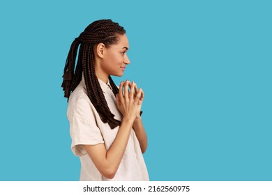 Side view of thoughtful woman with black dreadlocks schemes something, keeps fingers together, considers over cunning plans, wearing white shirt. Indoor studio shot isolated on blue background.