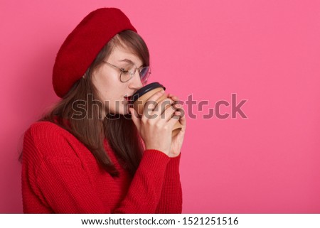 Side view of thoughtful young darkhaired woman with disposable cup of takeaway coffee, standing against pink background dressess sweater and red beret, copy space for advertisment or promotion.