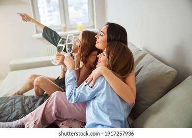 Side View Of Three Close Friends With Champagne Flutes Sitting On The Bed At Home