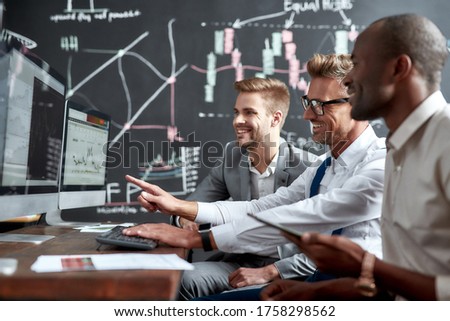Side view of three colleagues traders sitting by desks in front of computer monitors and discussing strategies while working in the office. Blackboard full of charts and data analyses in background