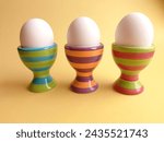 Side view of three white eggs in striped, colorful egg cups on the yellow background.