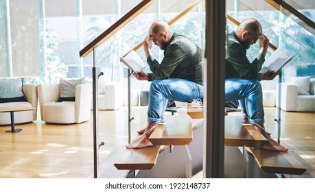 Side View Through Glass Of Pensive Focused Bearded Man In Casual Wear And Glasses Reading Book Pensively With Hand On Forehead While Sitting On Stairway In Penthouse Apartment